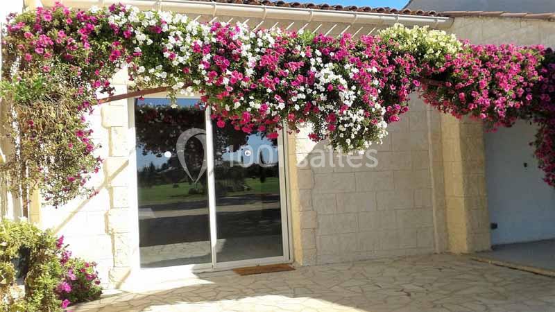 Pergola ornée de fleurs roses et blanches devant une maison en pierre avec une porte-fenêtre donnant sur un jardin.