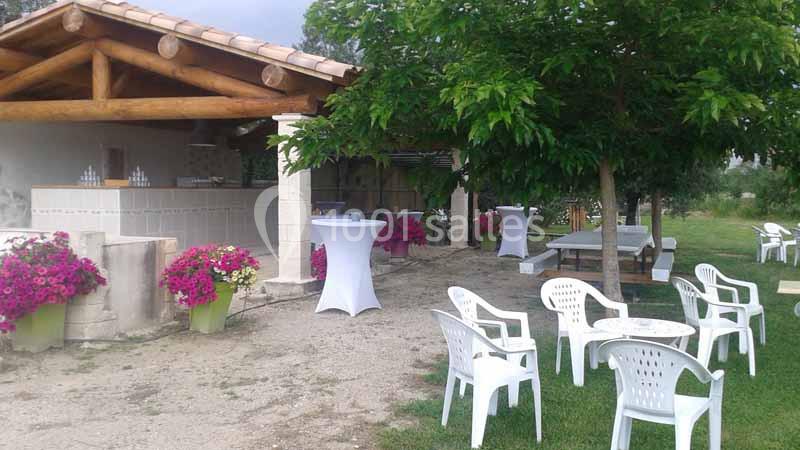 Espace extérieur avec bar en bois, tables et chaises blanches, fleurs colorées et arbres offrant de l'ombre.