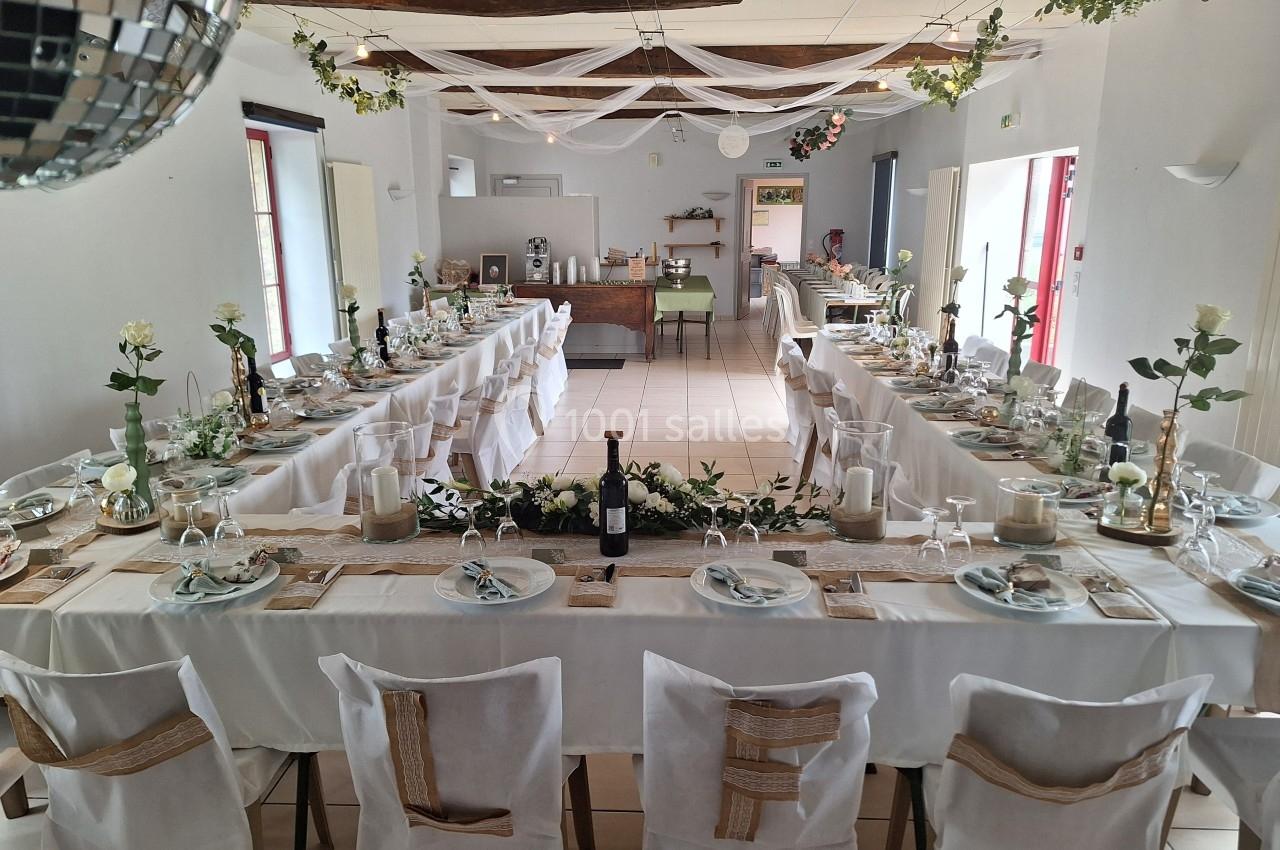 Salle décorée pour un repas festif avec longues tables dressées, nappes blanches, fleurs et guirlandes suspendues.
