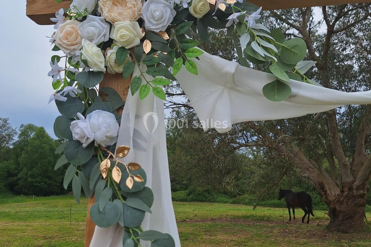 Arrangement floral blanc et vert avec des touches dorées, drapé de tissu blanc, fixé à une structure en bois dans un champ.
