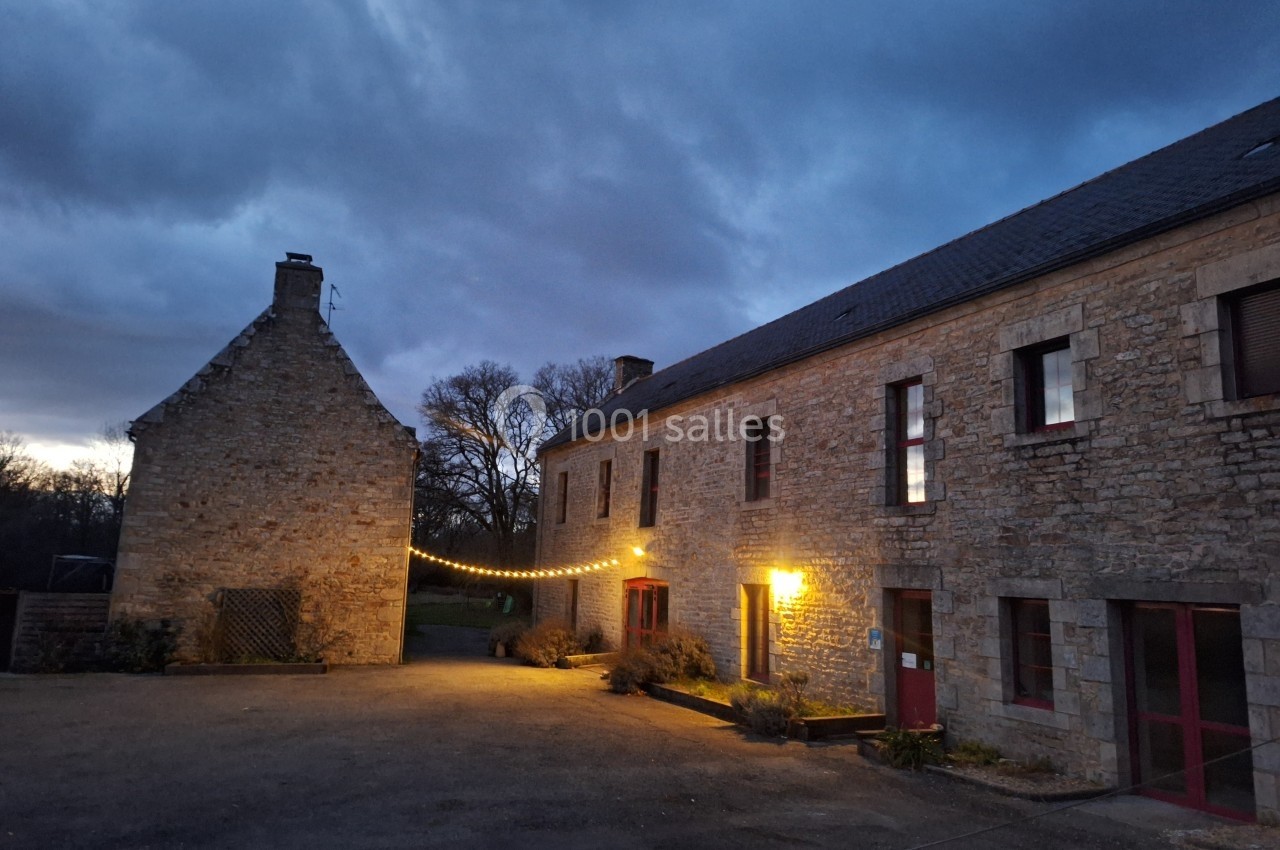 Cour d'une maison en pierre éclairée par des guirlandes lumineuses et des lampes, sous un ciel nuageux au crépuscule.