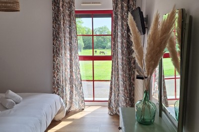 Chambre lumineuse avec lit simple, commode verte, miroir, vase de pampas et fenêtre donnant sur un paysage verdoyant.
