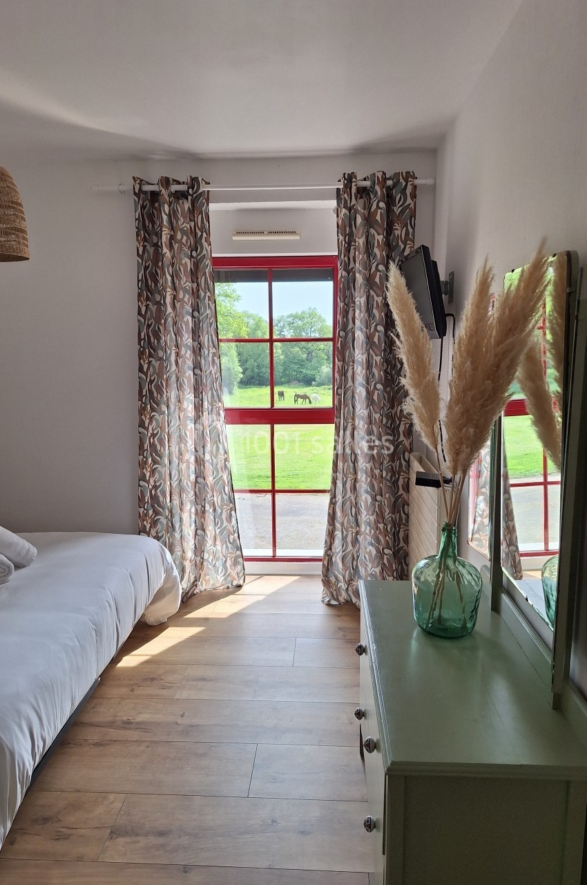 Chambre lumineuse avec lit simple, commode verte, miroir, vase de pampas et fenêtre donnant sur un paysage verdoyant.
