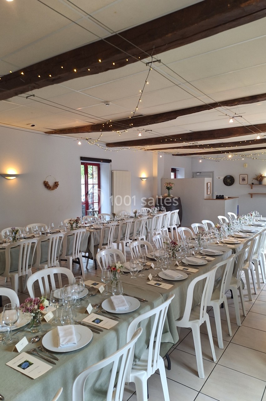 Salle décorée pour un repas festif avec longues tables dressées, nappes claires, vaisselle élégante et guirlandes lumineuses.