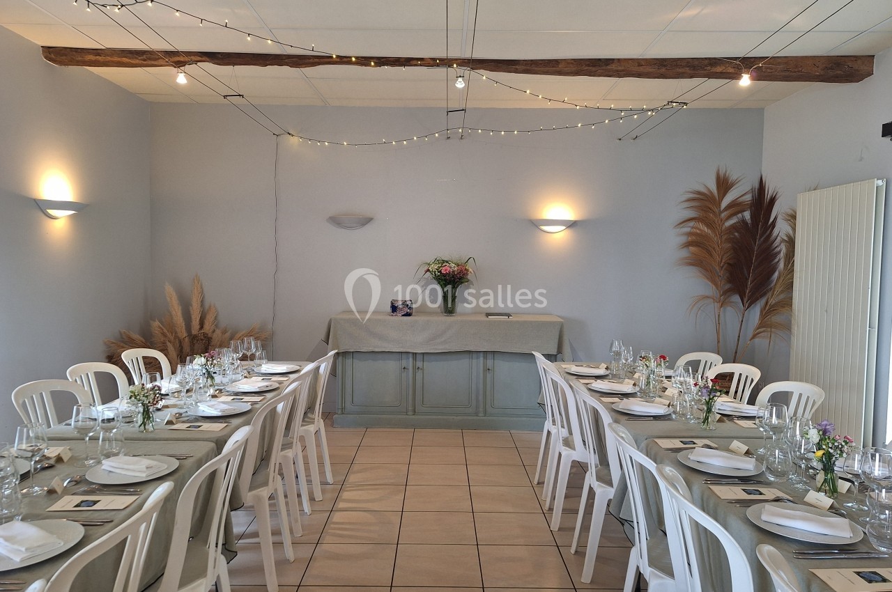 Salle décorée pour un repas, avec tables alignées, nappes beiges, chaises blanches et bouquet de fleurs sur un buffet.