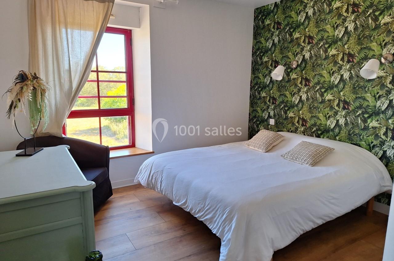 Chambre lumineuse avec un lit double, mur à motif végétal, fenêtre rouge et mobilier simple en bois.