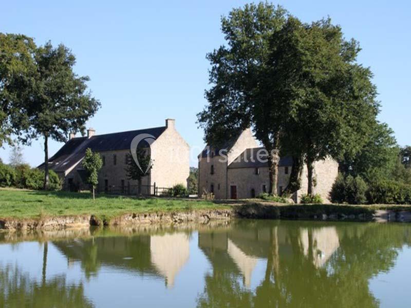 Deux maisons en pierre entourées d'arbres, reflétées dans un étang sous un ciel bleu.