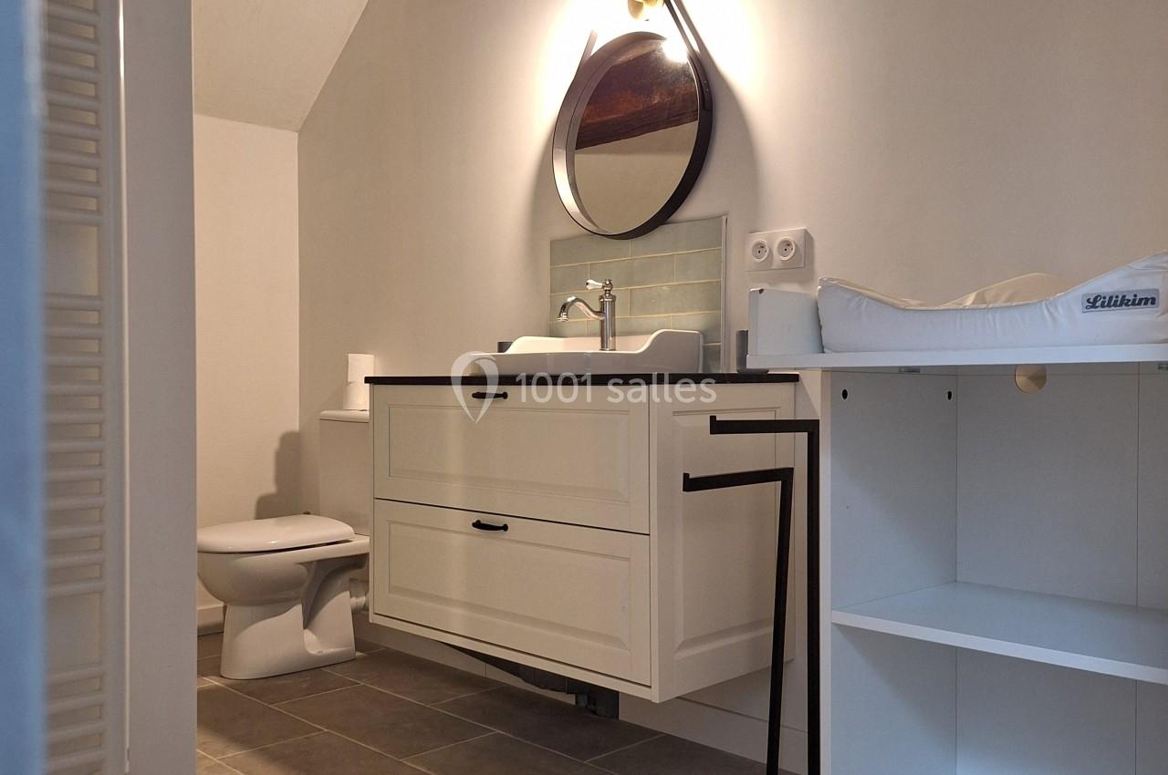 Salle de bain avec meuble blanc, lavabo, miroir mural triangulaire, table à langer et poutre apparente au plafond.
