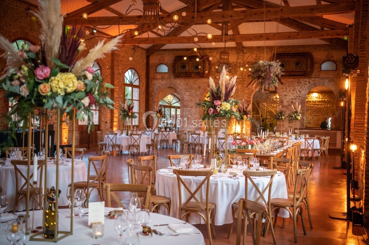 Salle de réception décorée avec des tables rondes, nappes blanches, chaises en bois et compositions florales élégantes.