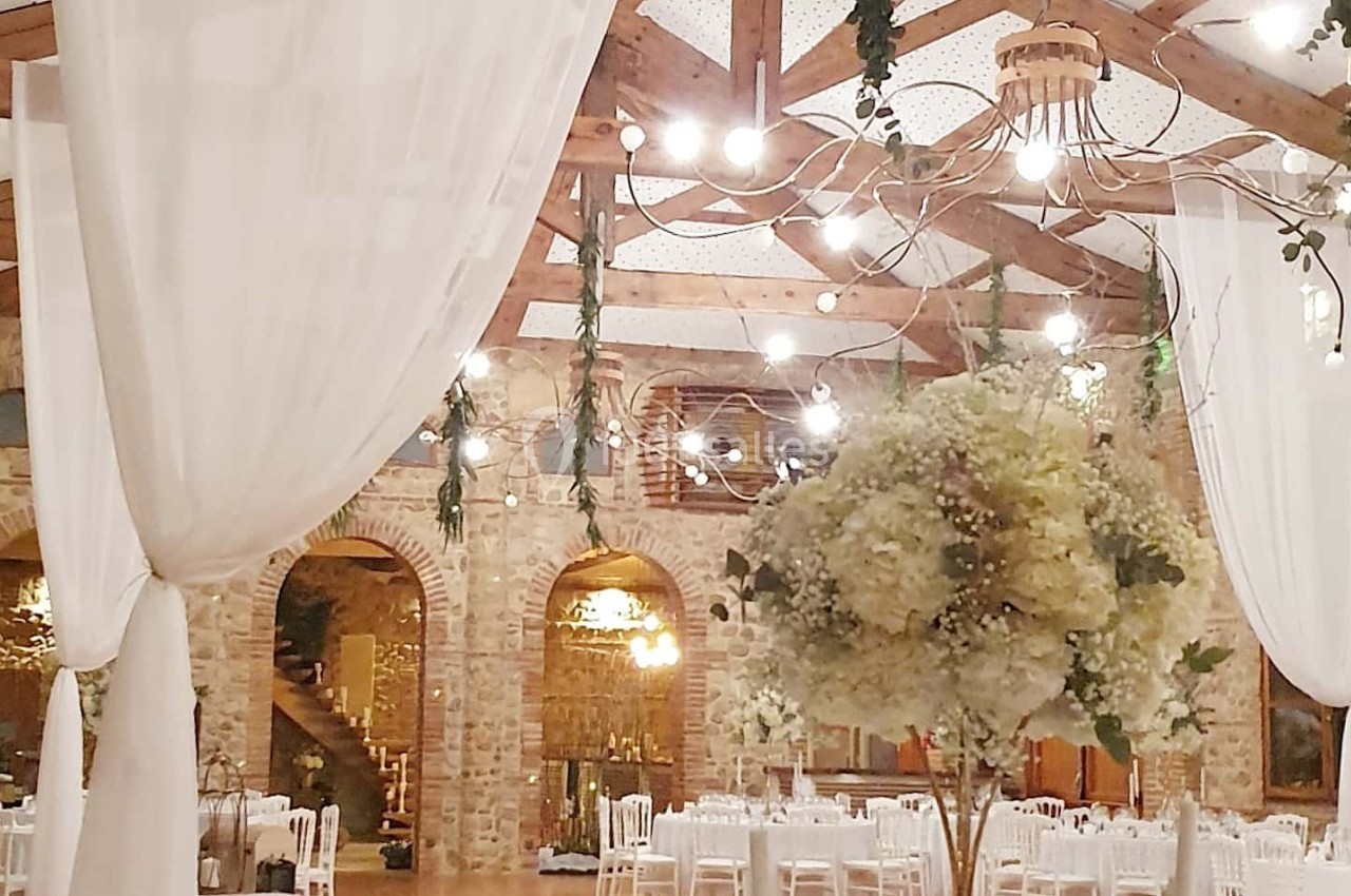 Salle de réception élégante avec poutres apparentes, lustres suspendus, rideaux blancs et tables décorées.