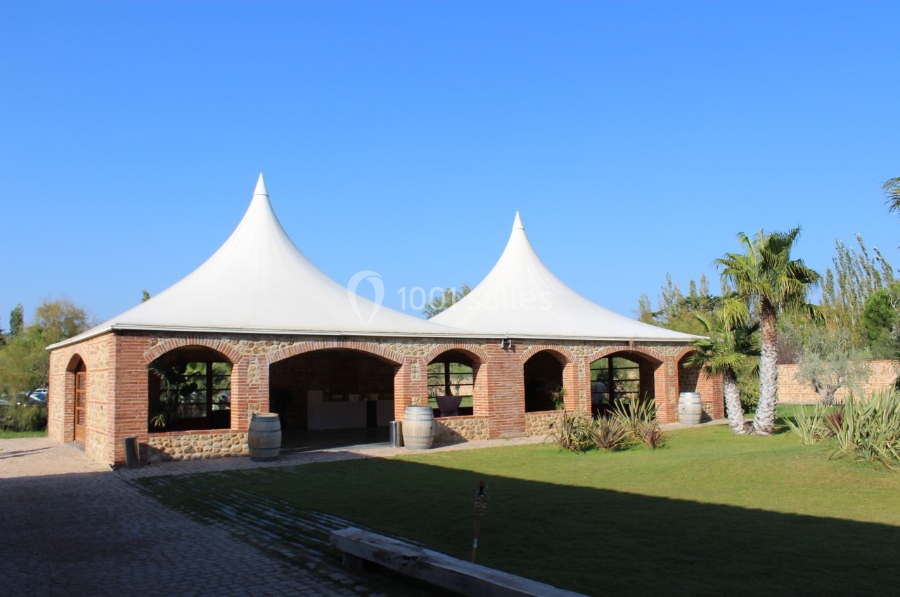 Structure en briques avec deux toits en toile blanche, entourée de pelouse, de palmiers et d'un ciel bleu clair.