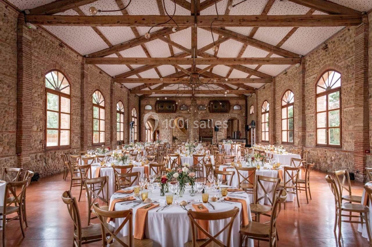 Salle de réception en briques avec poutres apparentes, tables rondes dressées et grandes fenêtres laissant entrer la lumière.