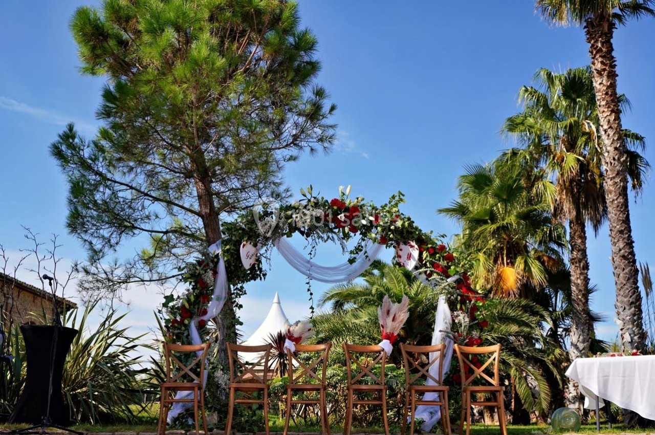 Arche fleurie entourée de chaises en bois dans un jardin ensoleillé avec des palmiers et un ciel bleu.