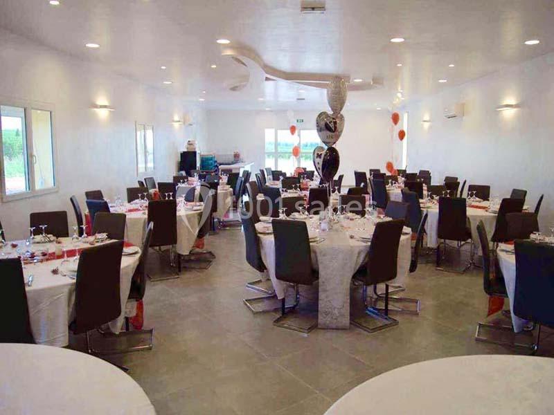 Salle de réception lumineuse avec tables rondes dressées, chaises noires et décorations en ballons.