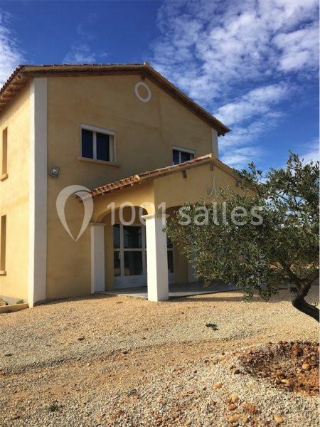 Façade d'une maison jaune à deux étages avec toit en tuiles, entourée de gravier et d'un olivier au premier plan.