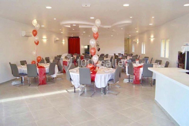 Salle de réception décorée avec des ballons rouges et blancs, tables rondes dressées avec nappes blanches et chaises grises.