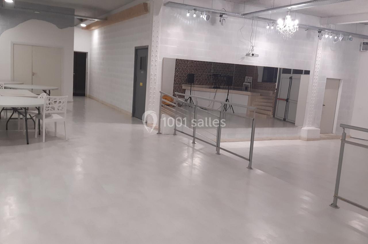 Salle lumineuse avec sol clair, tables, chaises blanches, miroir mural, rampe d'accès et éclairage suspendu.