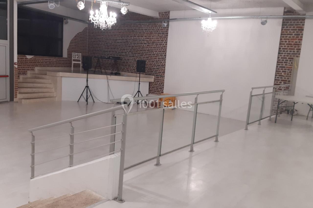 Salle vide avec sol clair, murs en briques, rampe d'accès, estrade avec enceintes et éclairage suspendu.