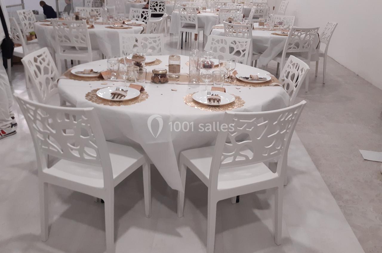 Salle de réception avec tables rondes dressées, nappes blanches, chaises blanches ajourées et décoration dorée.