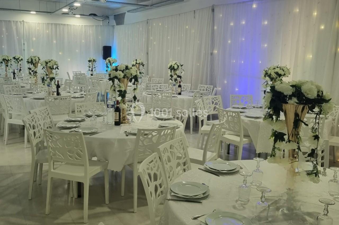 Salle de réception décorée avec des tables rondes, nappes blanches, chaises ajourées et centres de table fleuris.