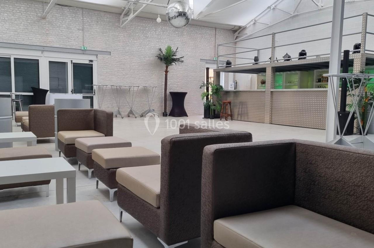 Salle lumineuse avec canapés, tables basses, bar moderne et décoration minimaliste, sous un plafond en verrière.