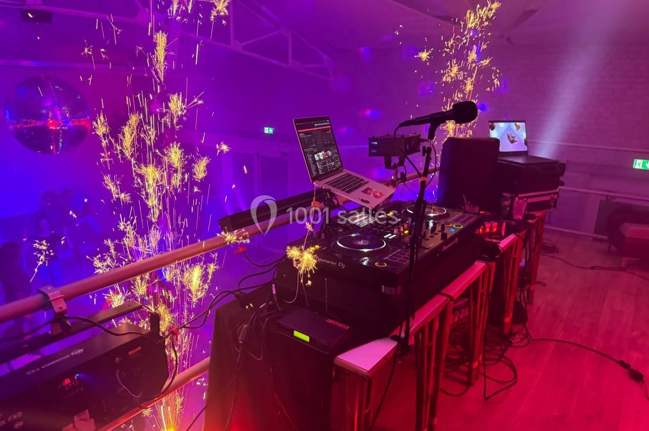 Poste de DJ avec platines et ordinateur, entouré d'effets pyrotechniques dans une salle éclairée par des lumières colorées.