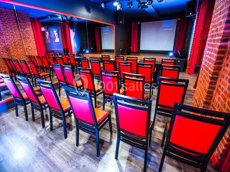 Salle avec des rangées de chaises rouges alignées face à un écran blanc, murs en briques et éclairage tamisé.