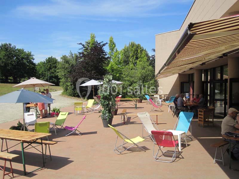 Terrasse ensoleillée avec chaises colorées, tables et parasols, entourée de verdure, quelques personnes assises et debout.