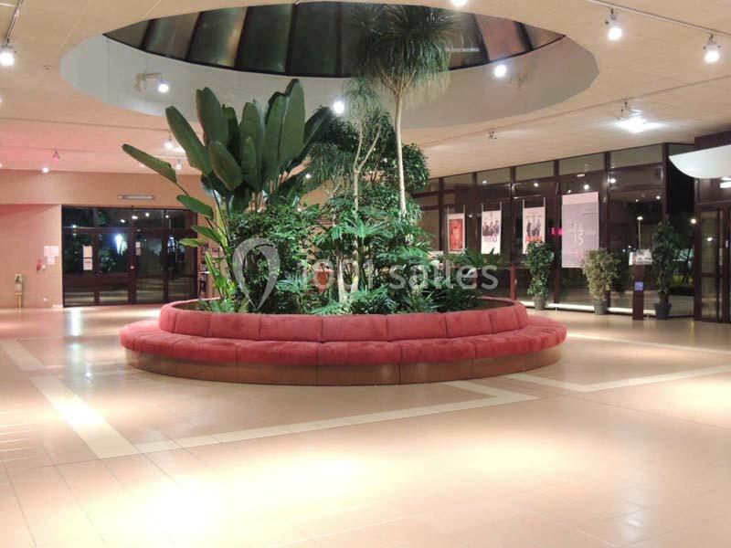 Un hall spacieux avec un grand canapé circulaire rouge entourant des plantes tropicales sous une verrière.