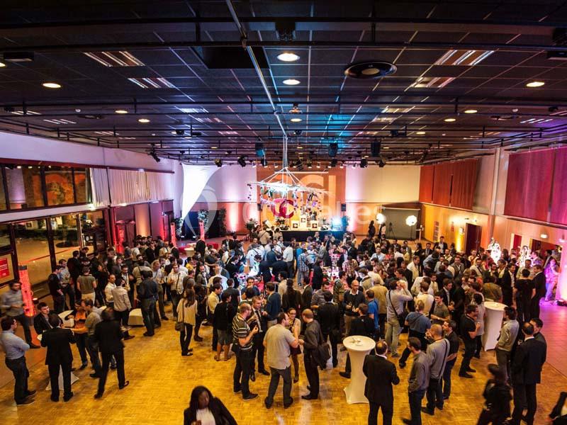 Salle de réception animée avec de nombreuses personnes debout, éclairage chaleureux et décorations suspendues.