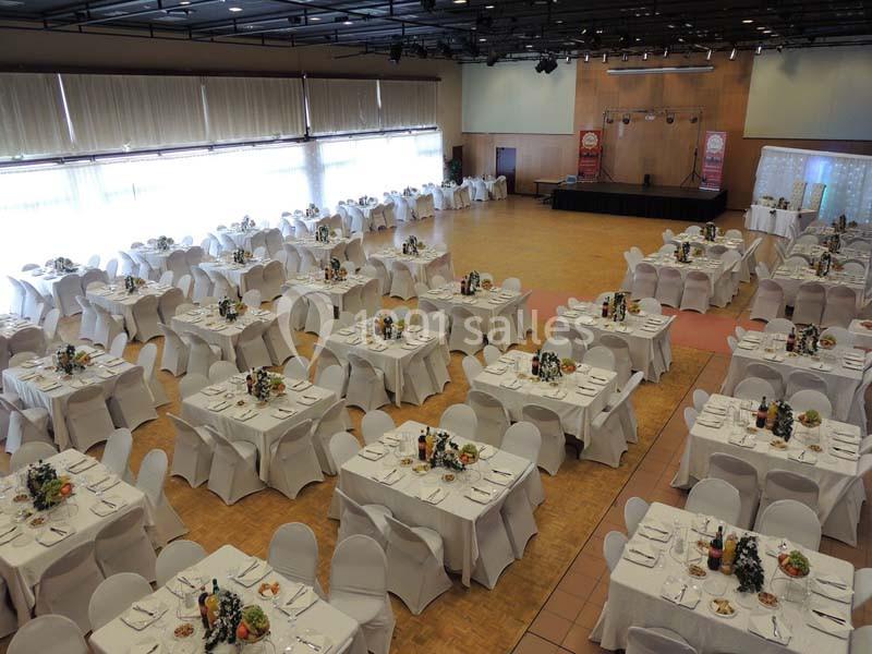 Salle de réception avec des tables rondes dressées, nappes blanches et décorations florales, prête pour un événement.