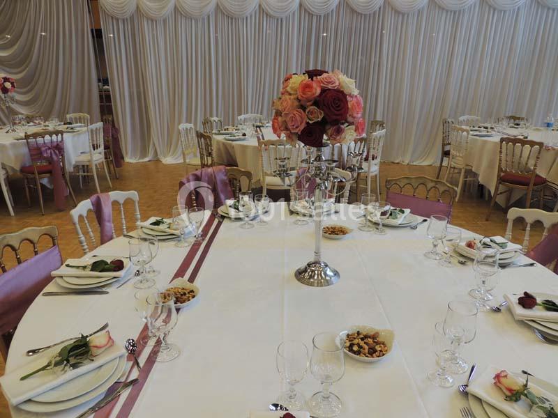Salle de réception décorée avec des tables rondes, nappes blanches, centres de table fleuris et chaises élégantes.