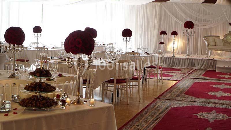 Salle de réception décorée avec des nappes blanches, des bouquets de fleurs rouges et des tapis rouges au sol.