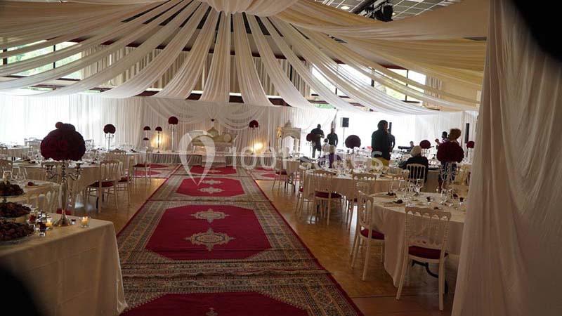 Salle de réception décorée avec des drapés blancs, tapis rouges et tables dressées pour un événement.