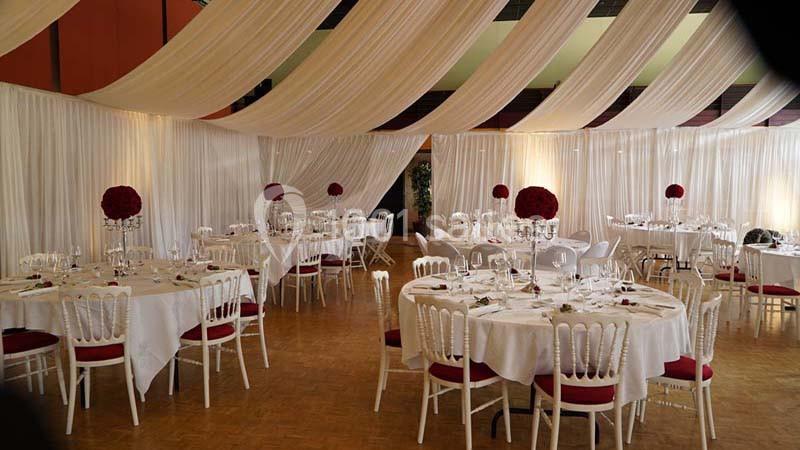 Salle de réception décorée avec des tables rondes, nappes blanches, chaises élégantes et centres de table fleuris rouges.