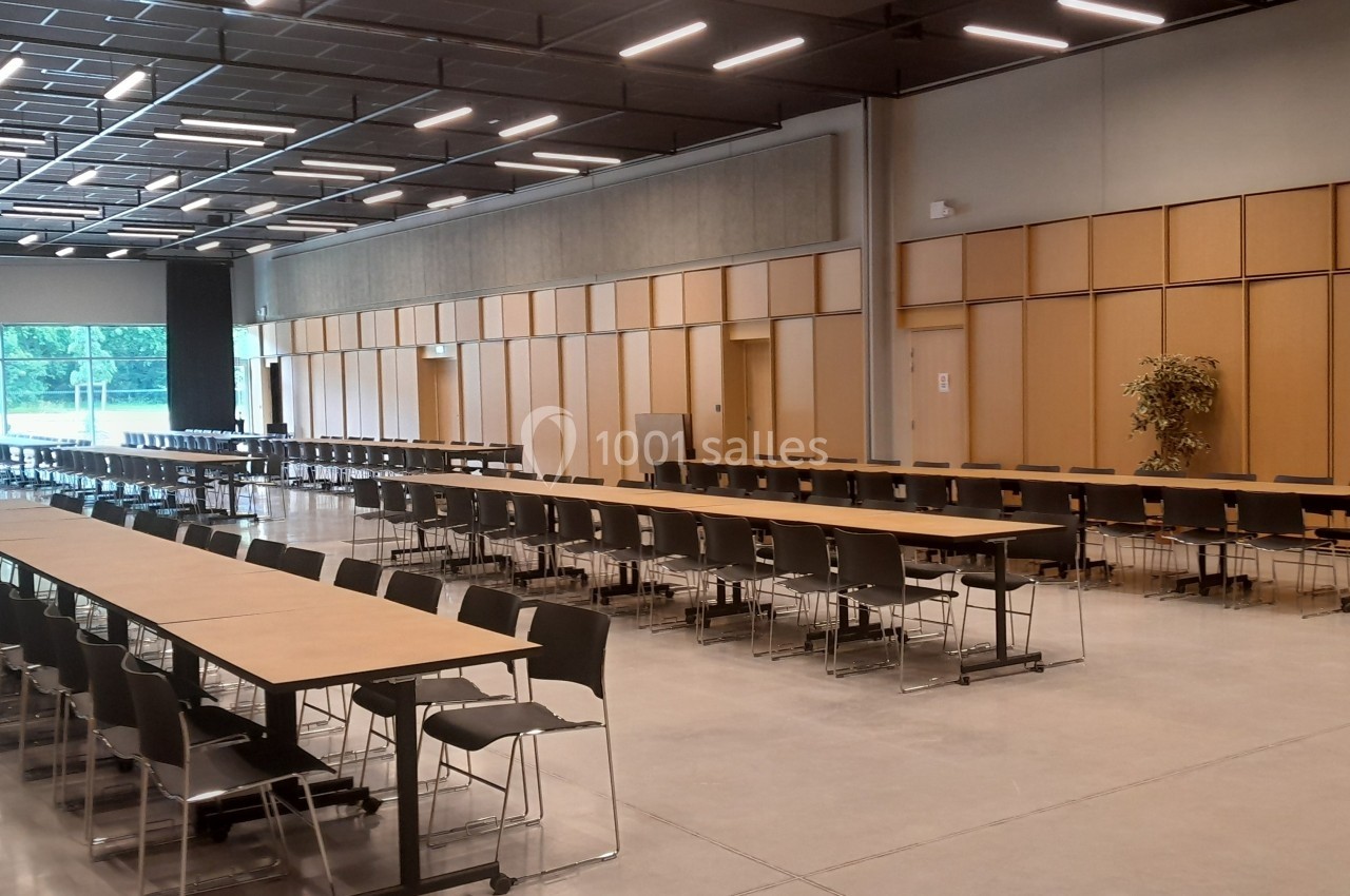 Salle spacieuse avec de longues tables en bois, des chaises noires alignées et un mur vitré laissant entrer la lumière…