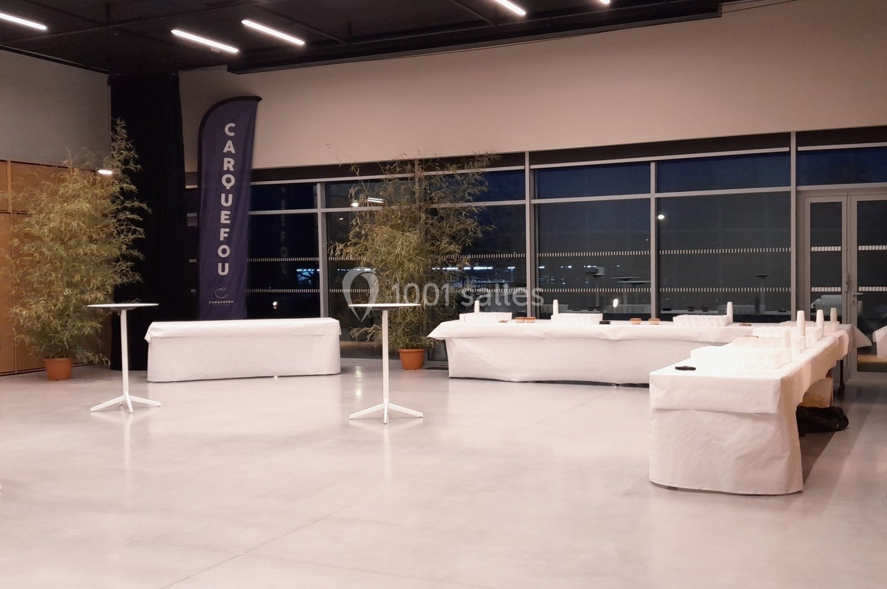 Salle de réception vide avec tables hautes et buffets recouverts de nappes blanches, entourée de baies vitrées et de plantes.