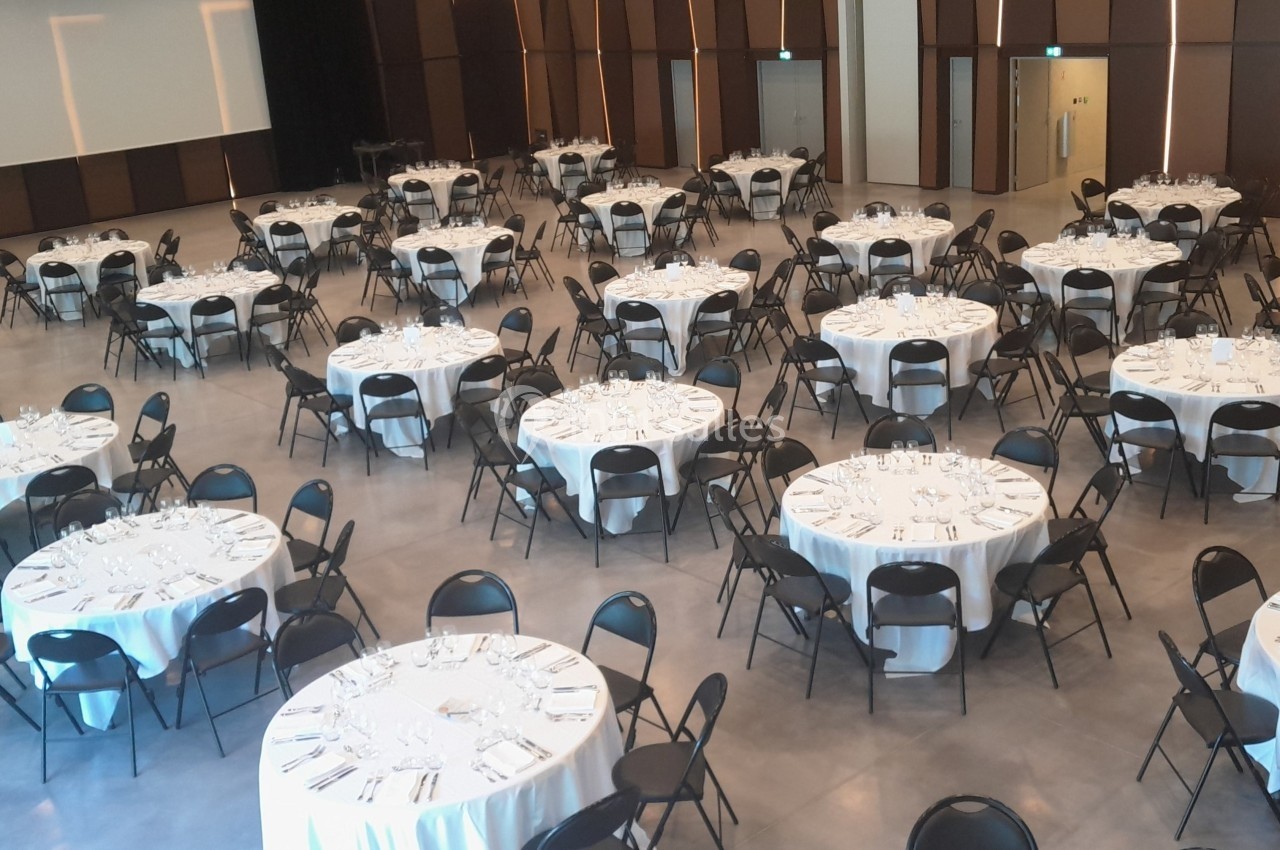 Salle de réception avec des tables rondes dressées, nappes blanches et chaises noires, prête pour un événement.