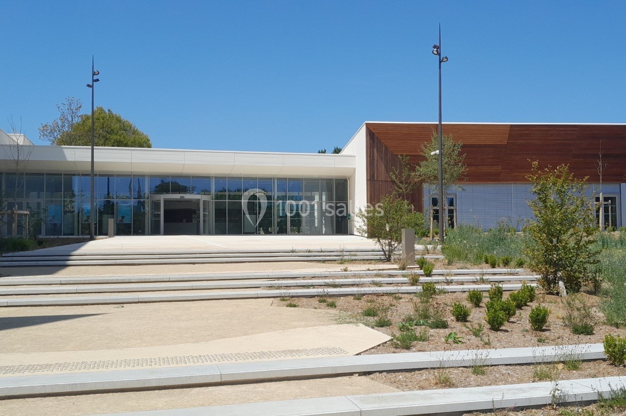 Entrée d'un bâtiment moderne avec façade vitrée et revêtement en bois, entourée de végétation et d'allées pavées.