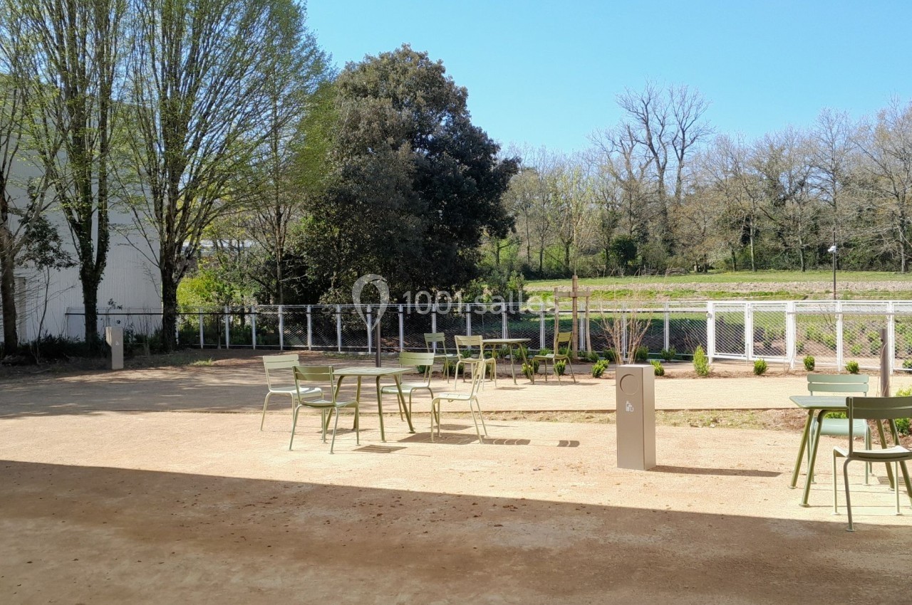 Espace extérieur avec tables et chaises sur sol en gravier, entouré de végétation et d'une clôture.