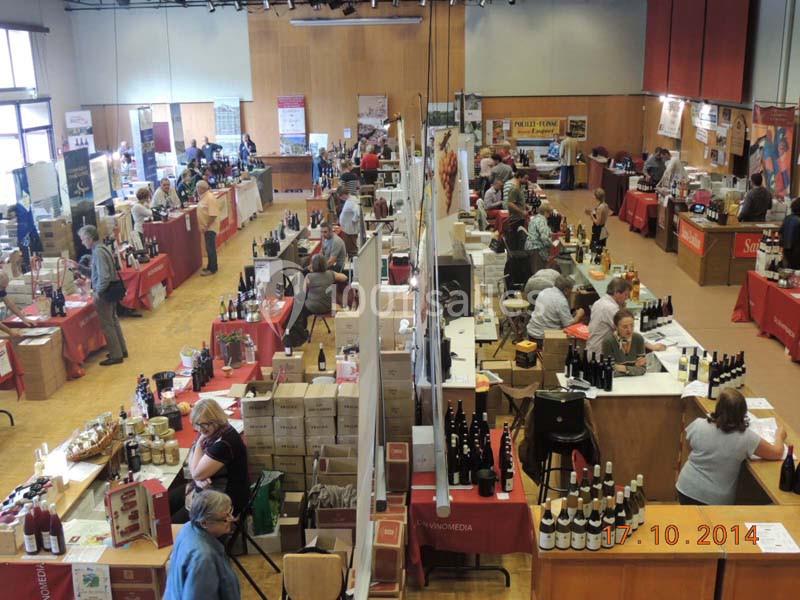 Vue d'ensemble d'un salon avec des stands proposant des produits, notamment des bouteilles de vin, dans une salle animée.