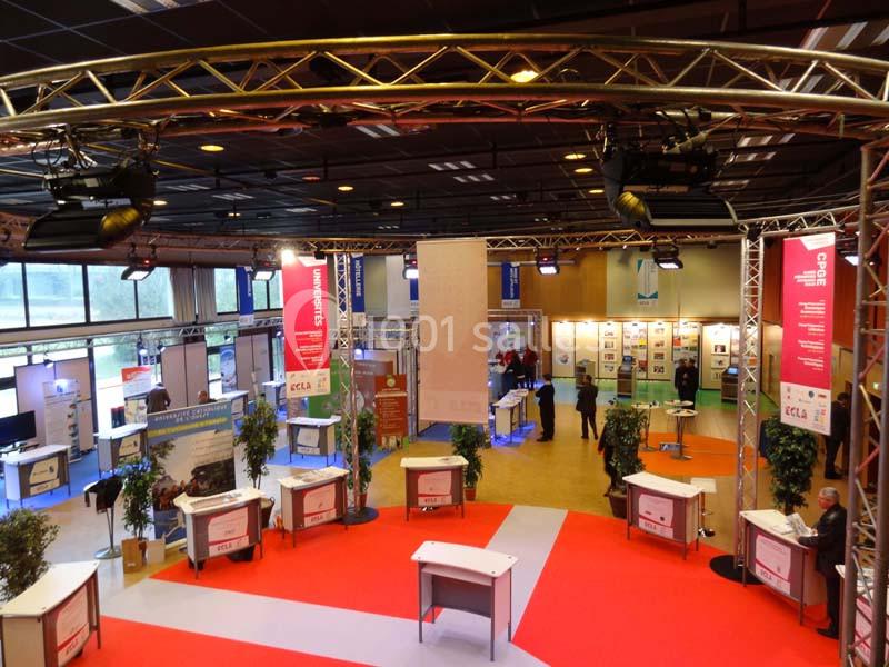 Vue d'un salon professionnel avec stands, éclairage suspendu et allées recouvertes de moquette rouge.