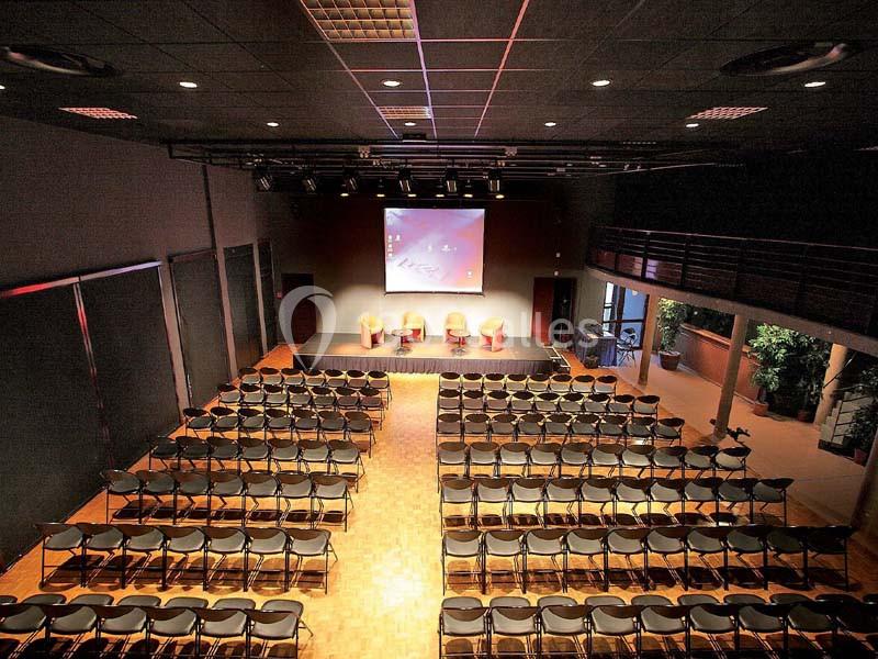 Salle de conférence avec rangées de chaises alignées, une scène équipée de fauteuils et un écran de projection.