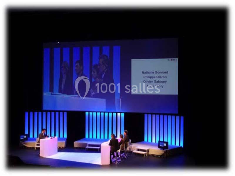 Des intervenants discutent sur une scène éclairée, avec un écran affichant des noms en arrière-plan.