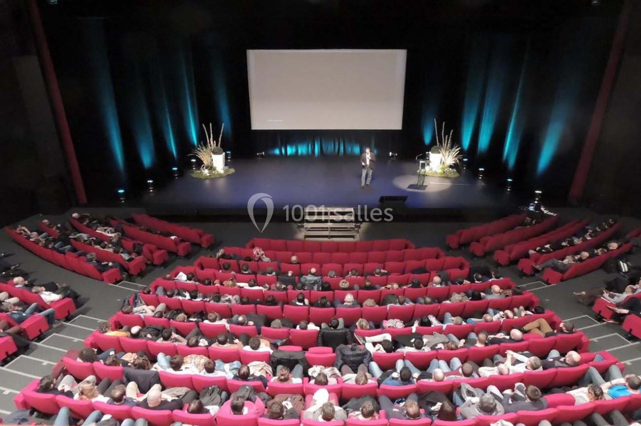 Salle de conférence avec un orateur sur scène, écran de projection et public assis dans des fauteuils rouges.