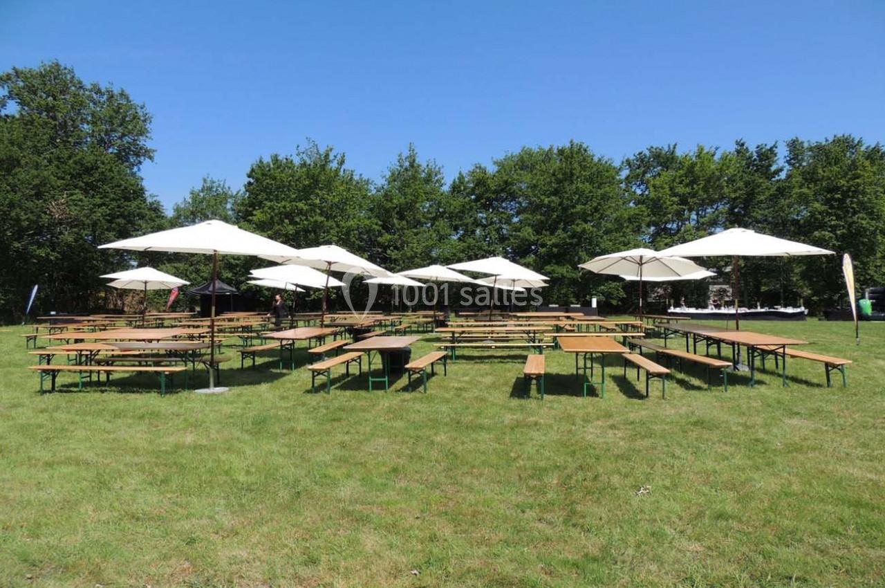 Tables et bancs en bois avec parasols blancs disposés sur une pelouse, entourés d'arbres sous un ciel dégagé.