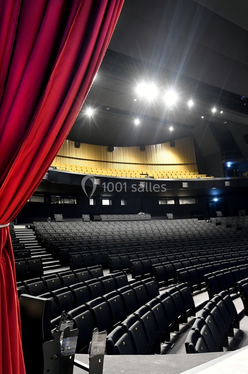 Salle de théâtre vide avec des sièges noirs, rideau rouge ouvert et éclairage de scène allumé.