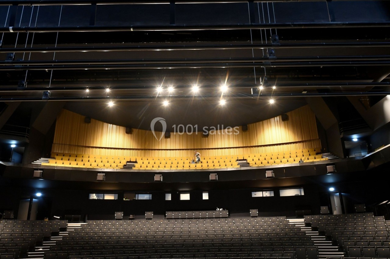 Salle de théâtre vide avec des rangées de sièges noirs et un balcon éclairé par des projecteurs.