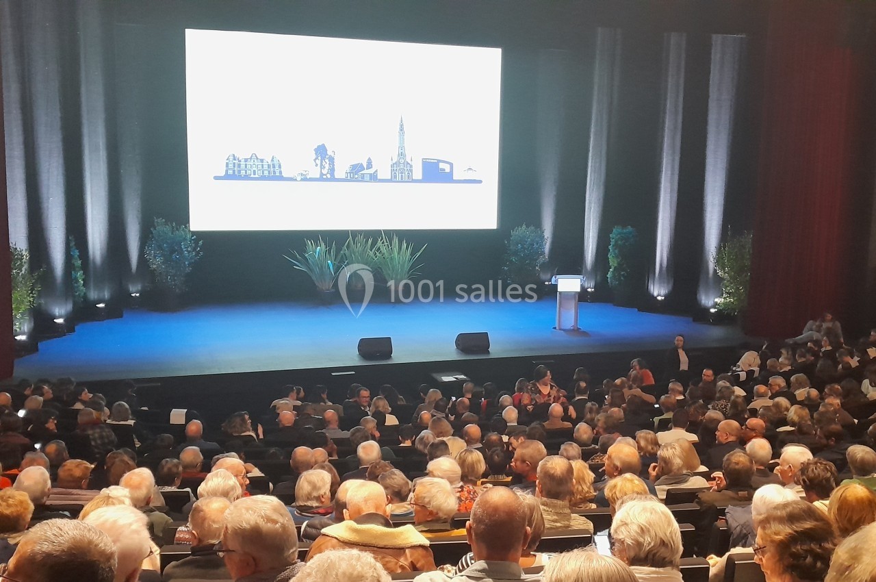 Salle de conférence remplie avec un public assis, scène éclairée avec écran et pupitre au centre.