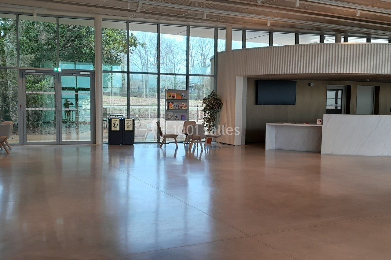 Hall d'entrée spacieux avec sol en béton, comptoir d'accueil, chaises, grandes baies vitrées et vue sur l'extérieur.